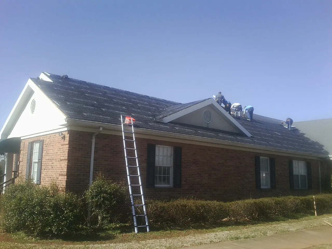 Crew tearing shingles off a brick ranch roof with a ladder set — Emergency Roof Repair work by our crew in Baxter Springs