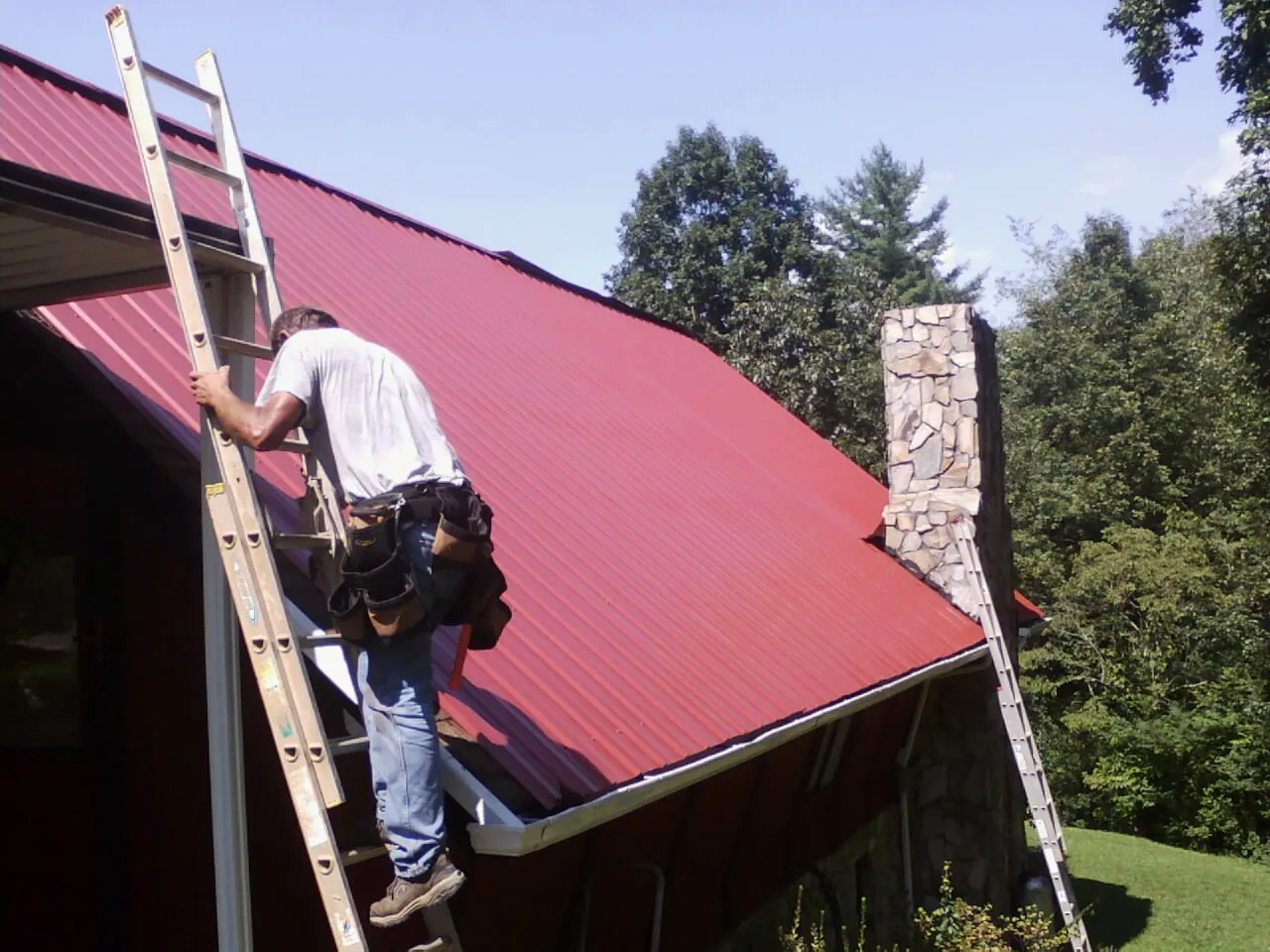 Roofer on a ladder beside a red metal roof and stone chimney — Soffit & Fascia Repair by Roofing Brothers in Baxter Springs